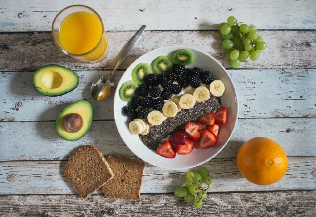 orange juice, grapes, avocado, green grapes, toast, and an orange surrounding a bowl of oats with fruit and chia seeds on top.